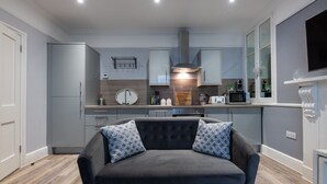 A modern kitchen with grey cabinets, a built-in oven, and a coffee machine. There is a dark grey sofa with patterned cushions in the foreground. - 2 Sussex Place (Plymouth)