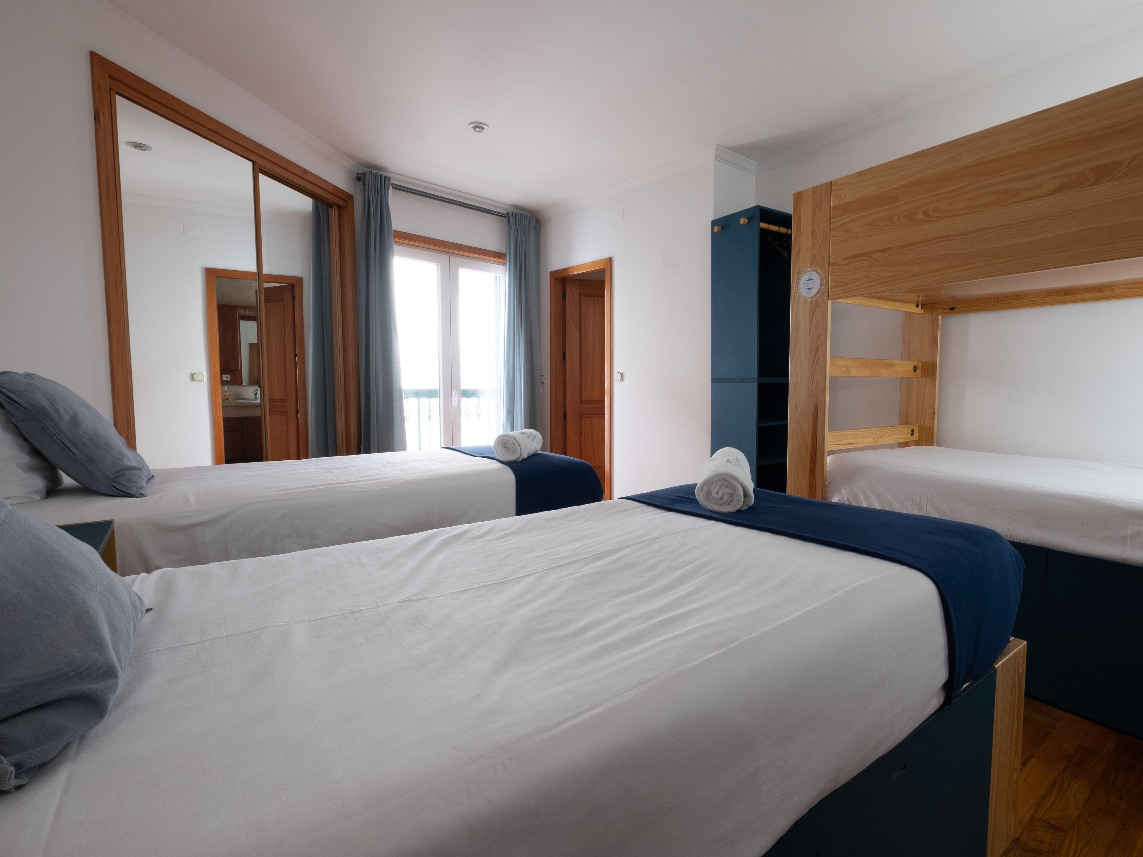 A hotel room with two single beds, a wooden wardrobe, a mirror, and a window with curtains.