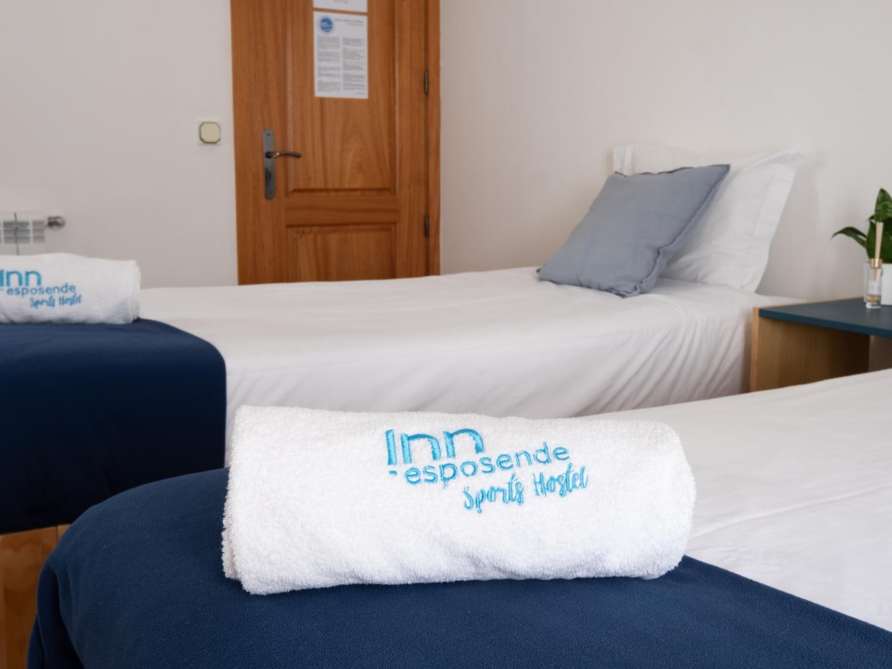 A hotel room with two beds, each with a white blanket and a towel with 'Inn esposende Sports Hostel' text. There is a wooden door and a small table with a plant.
