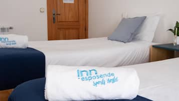 A hotel room with two beds, each with a white blanket and a towel with 'Inn esposende Sports Hostel' text. There is a wooden door and a small table with a plant.