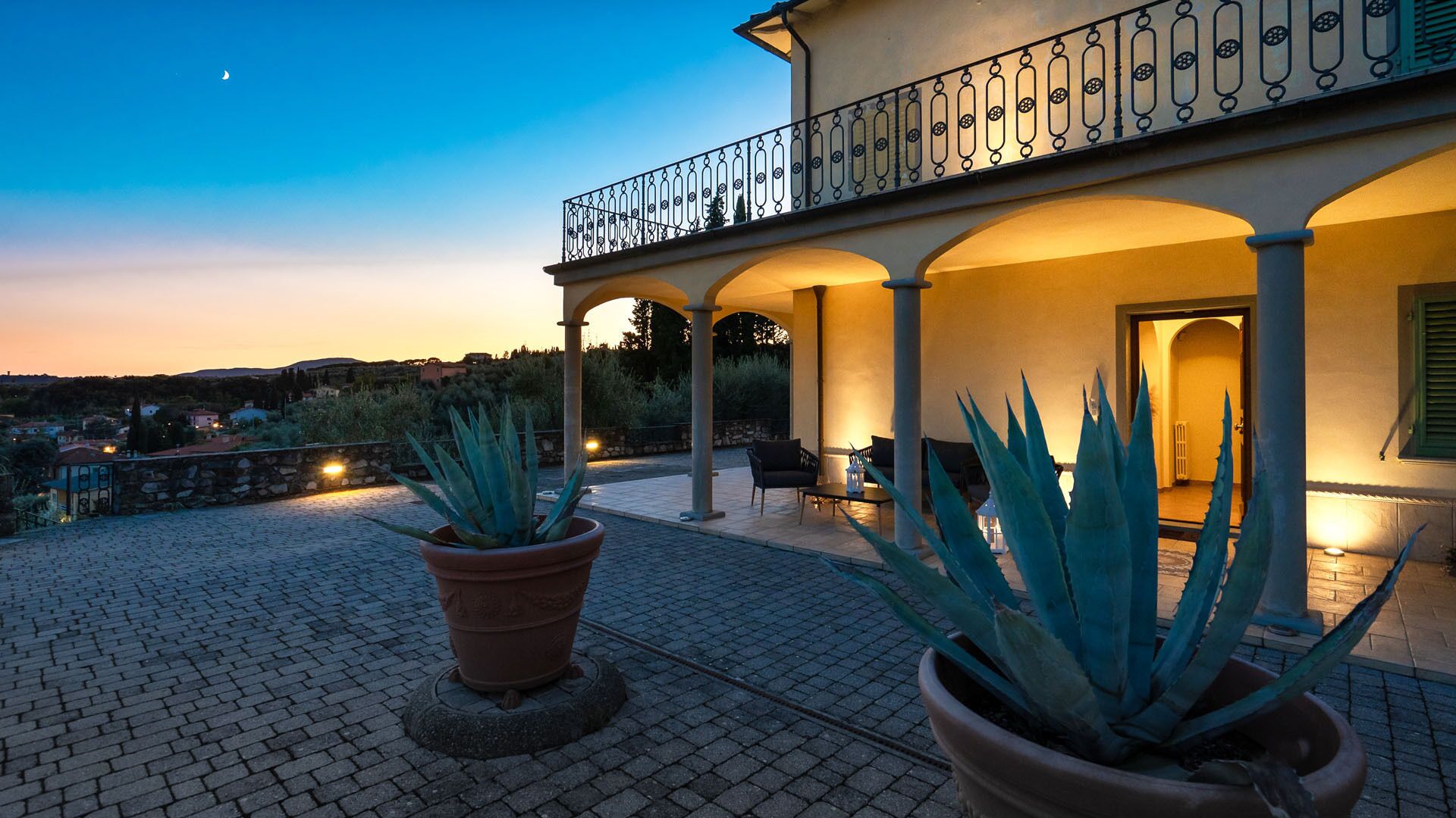 A terrace with potted agave plants, a balcony with ornate railing, and a view of the surrounding landscape during twilight.