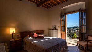 A bedroom with a bed, a nightstand, a lamp, a radiator, and a view of a city through an open door. - VILLA IL TORRIONE: Elegance and luxury home between Lucca and Florence with SPA (Pescia)