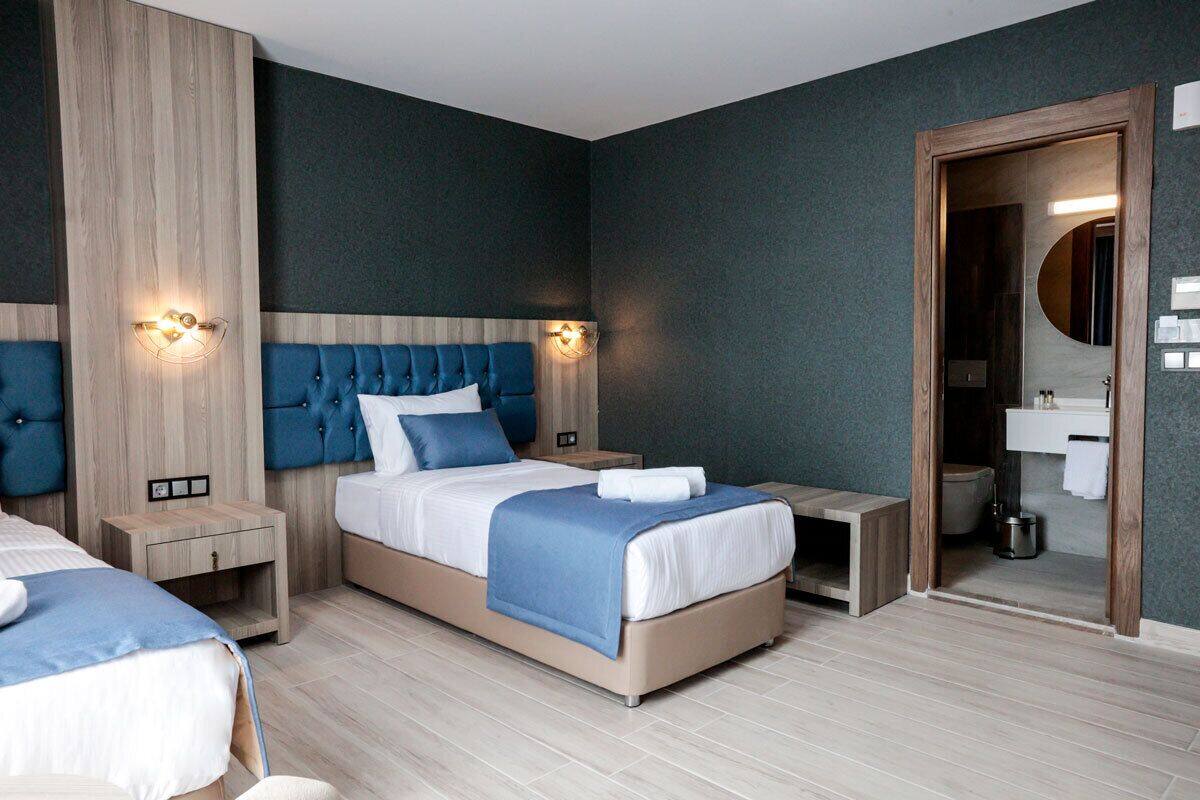 Una habitación de hotel moderna con una cama grande, una cama más pequeña, una mesita de noche y un cabecero de madera, además de un baño visible a través de una puerta abierta.