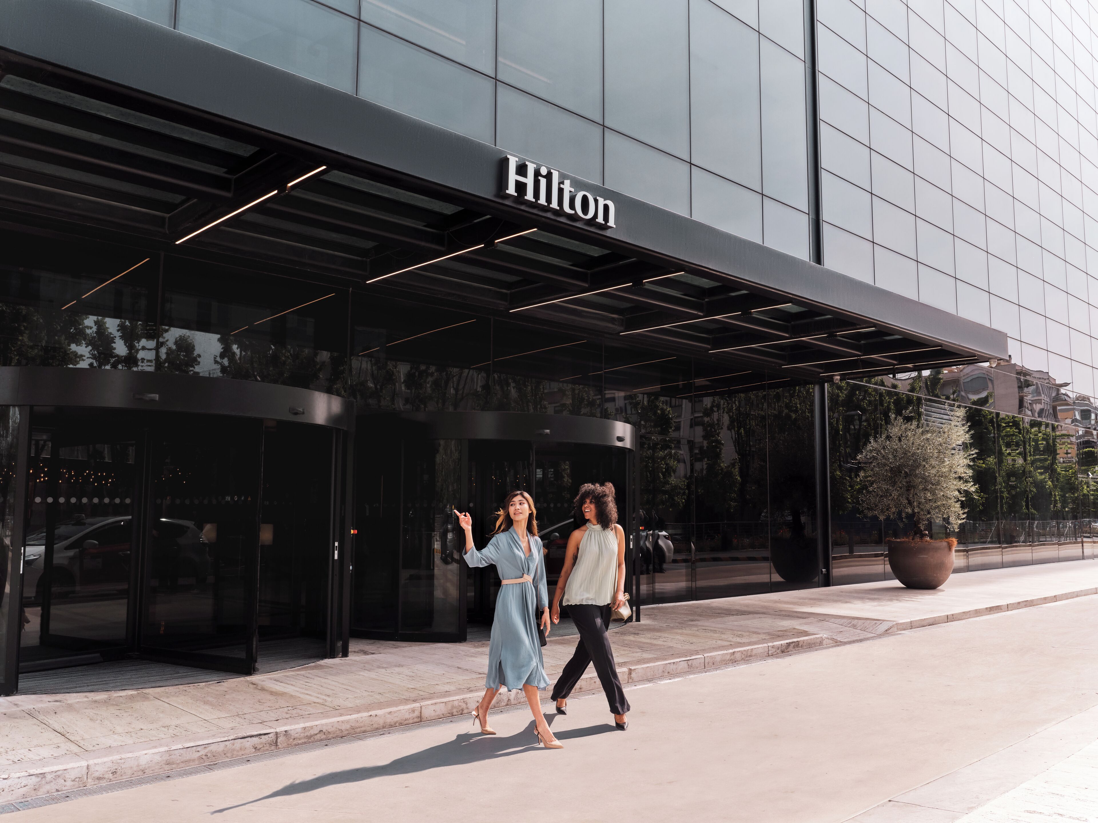 Exterior de um hotel Hilton com fachada de vidro moderna, uma planta grande em vaso e uma entrada ampla e convidativa.