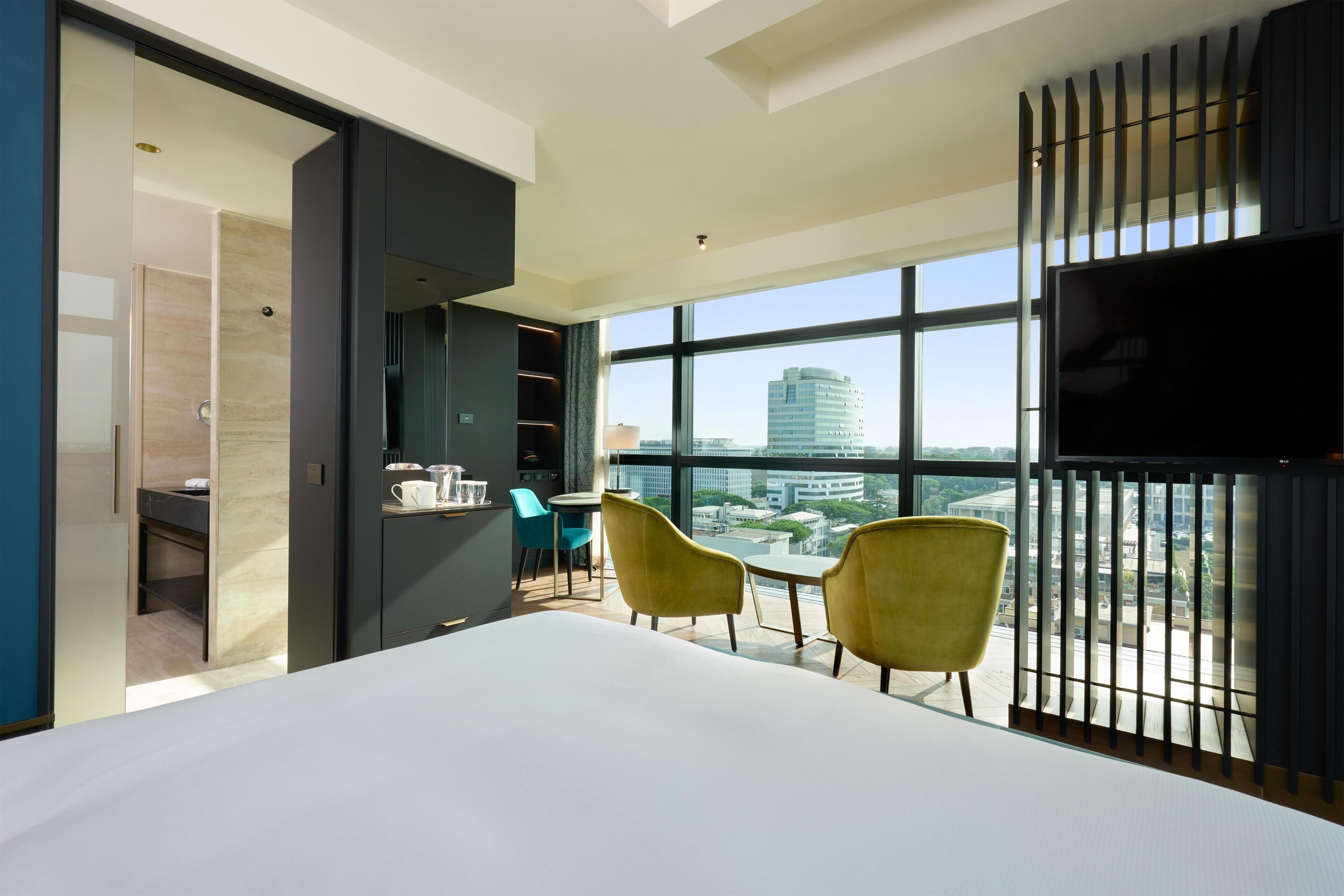 A modern hotel room with a large bed, a dining area with chairs, a TV, and a view of the cityscape through large windows.