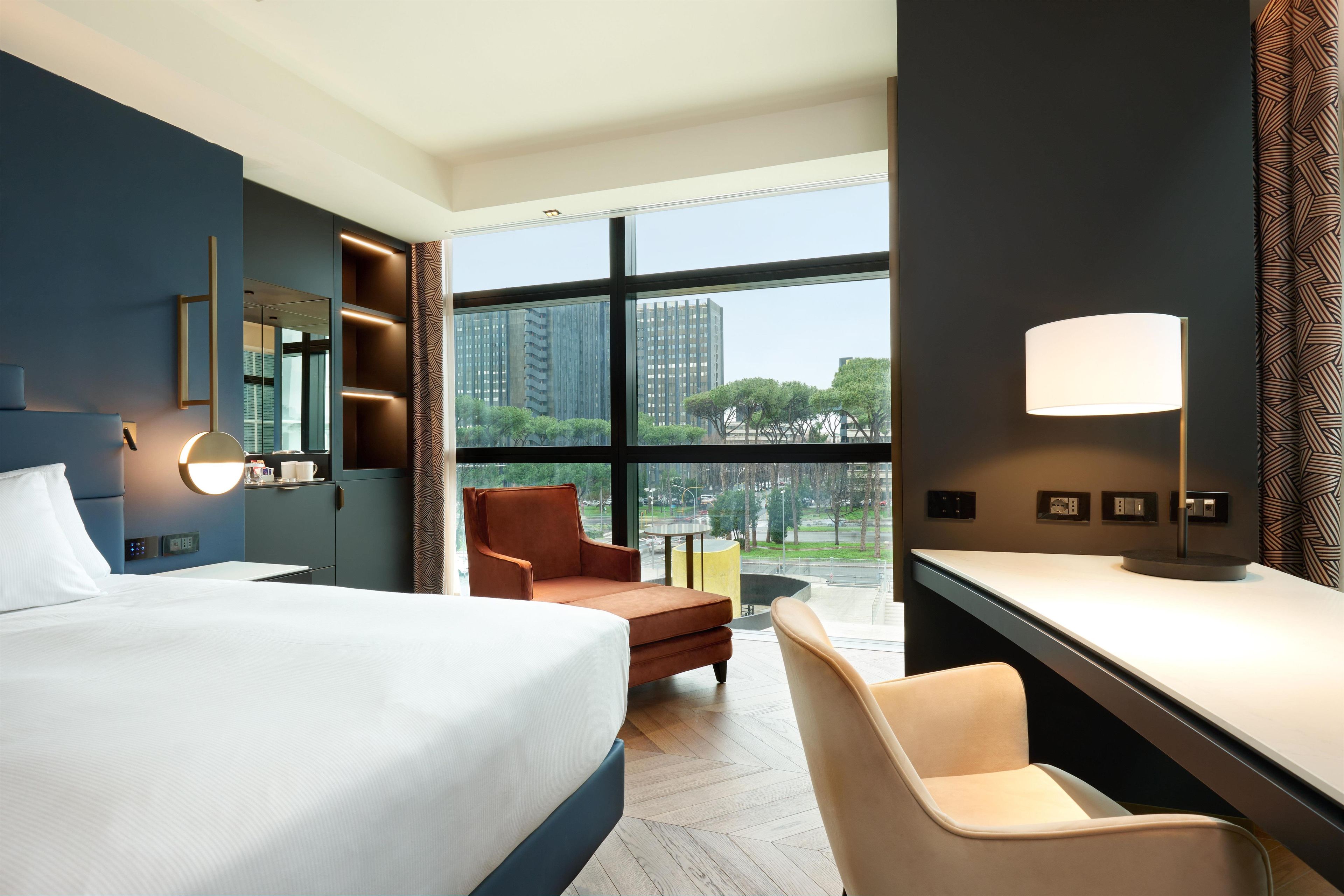 Une chambre d’hôtel moderne dotée d’un grand lit, d’un bureau, d’une chaise et offrant une vue sur le paysage urbain grâce à de grandes fenêtres.