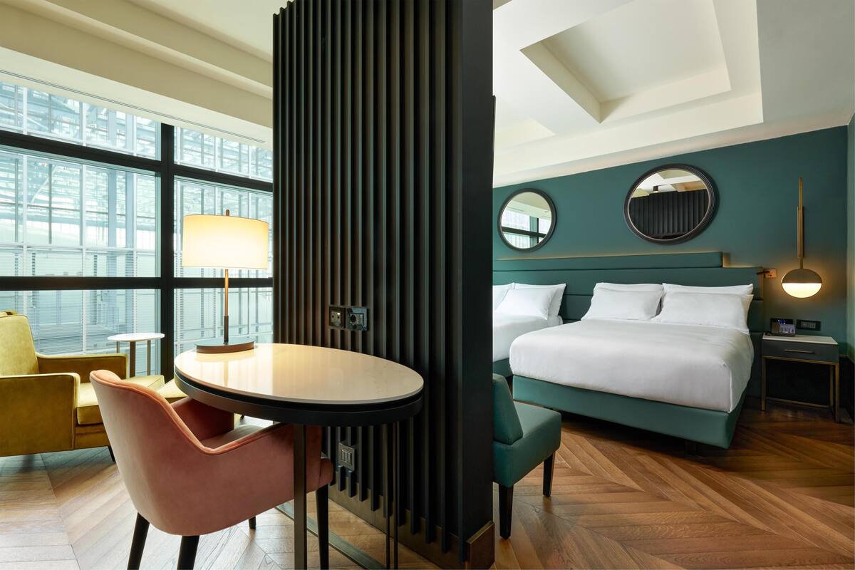 A hotel room with a bed, a round mirror, a bedside table, a lamp, a chair, and a view of the city through large windows.