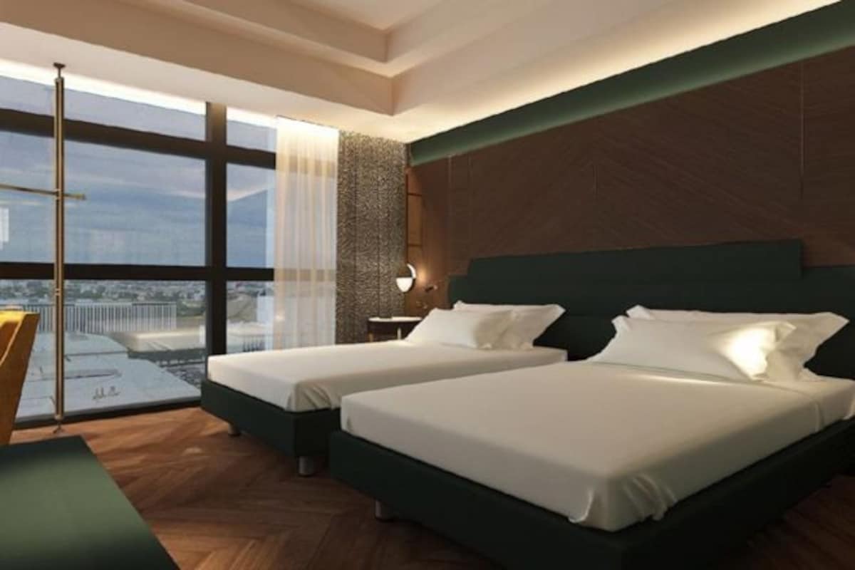 A modern hotel room with two beds, a large window with a city view, and a wooden floor.