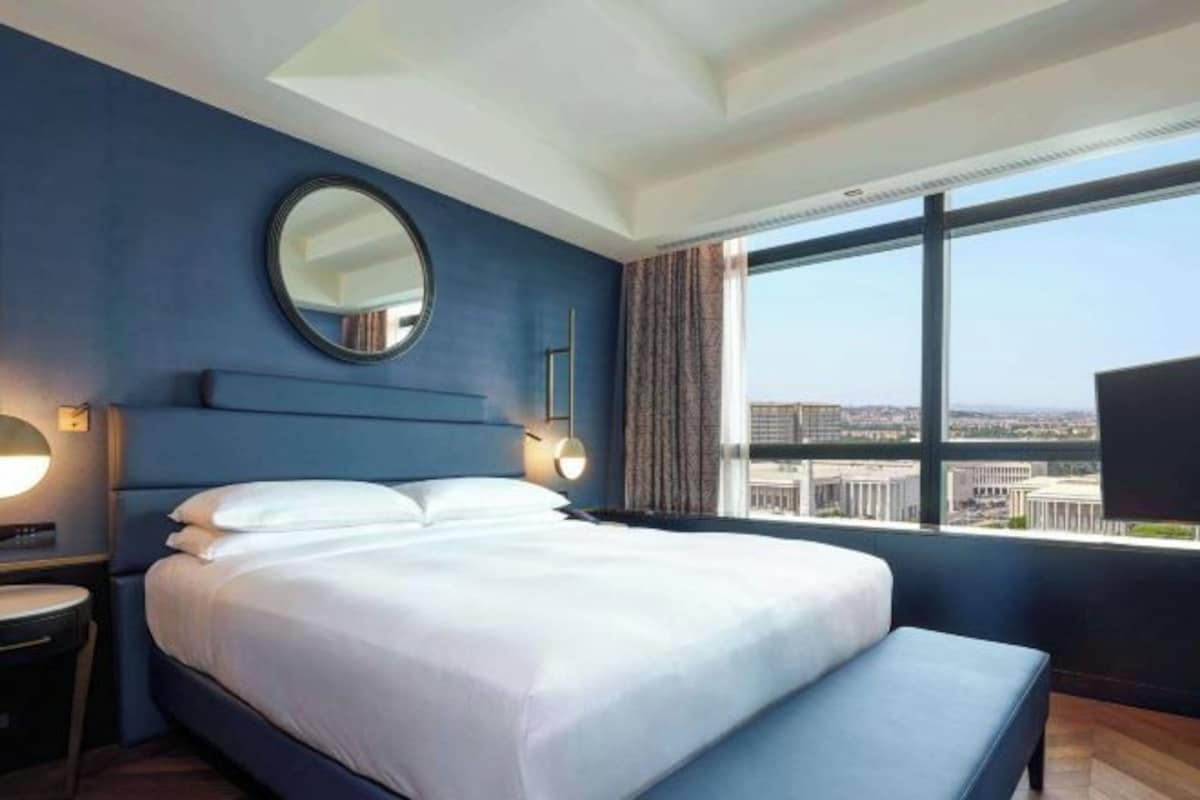 A hotel room with a large bed, a round mirror, a bedside table, and a view of the city through a large window.