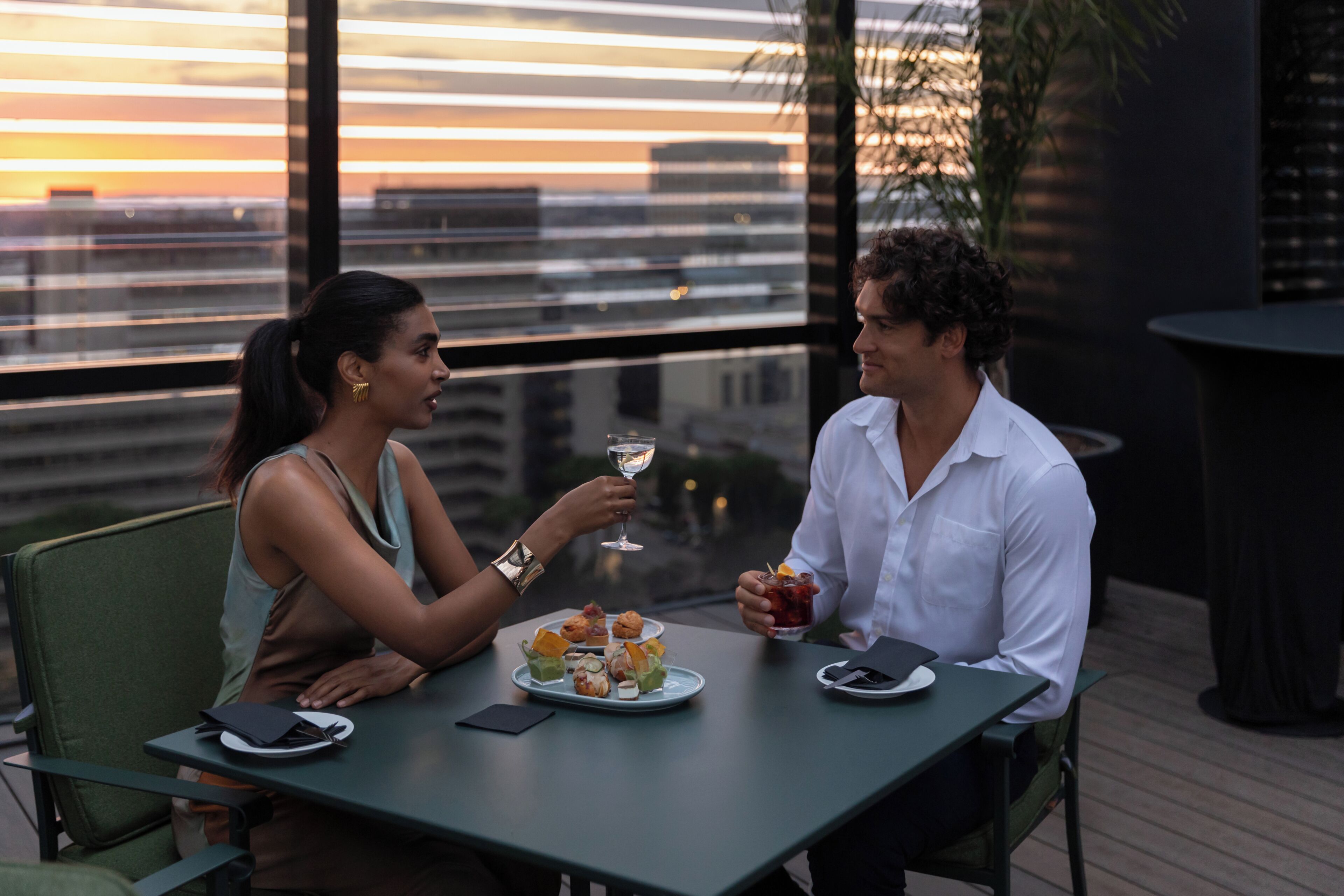 A couple dining at an outdoor table with a city view, plates of food, and drinks.