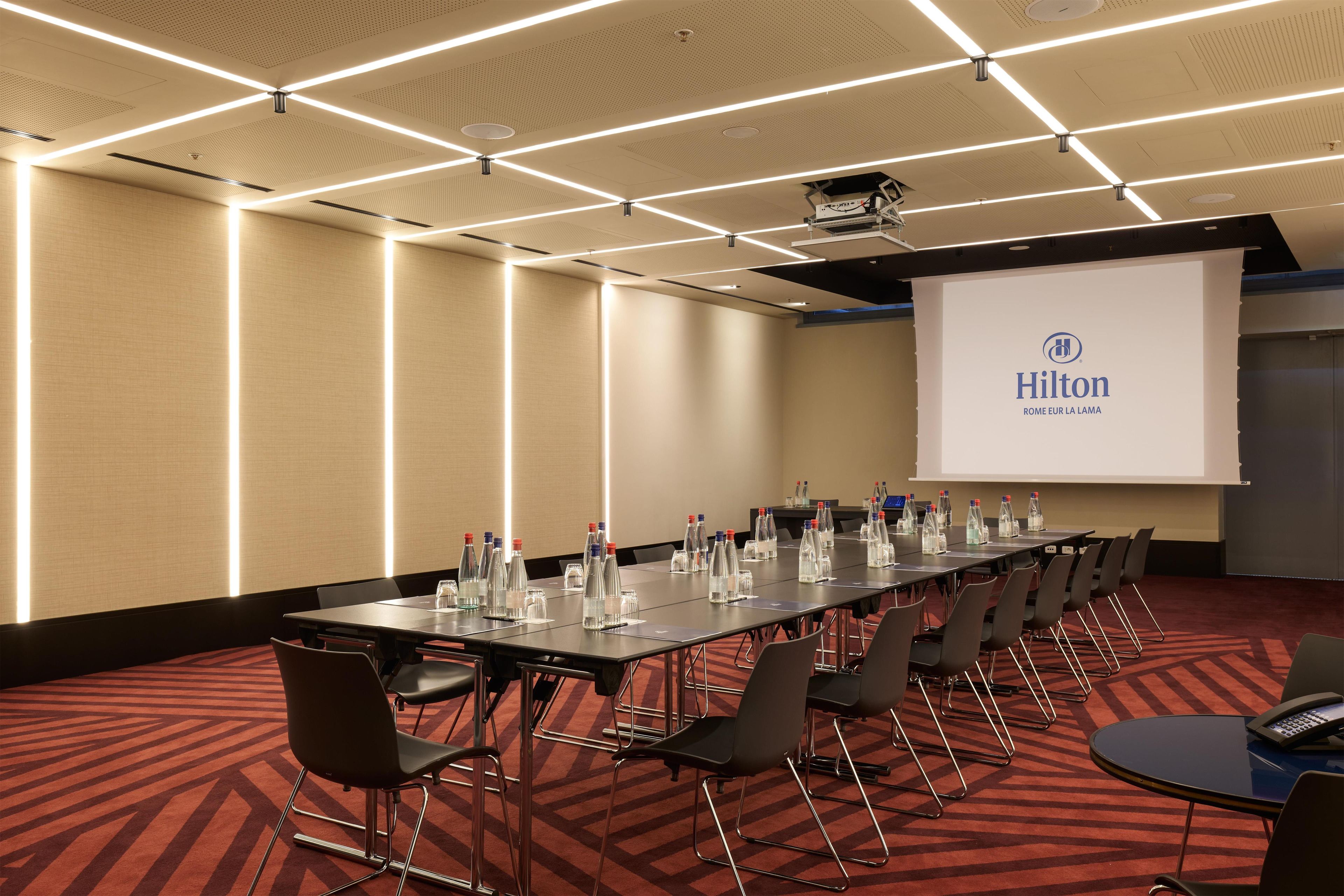 Ein Konferenzraum mit einem langen Tisch, Stühlen, Wasserflaschen und einer Projektionsleinwand, auf der das Hilton-Logo zu sehen ist.