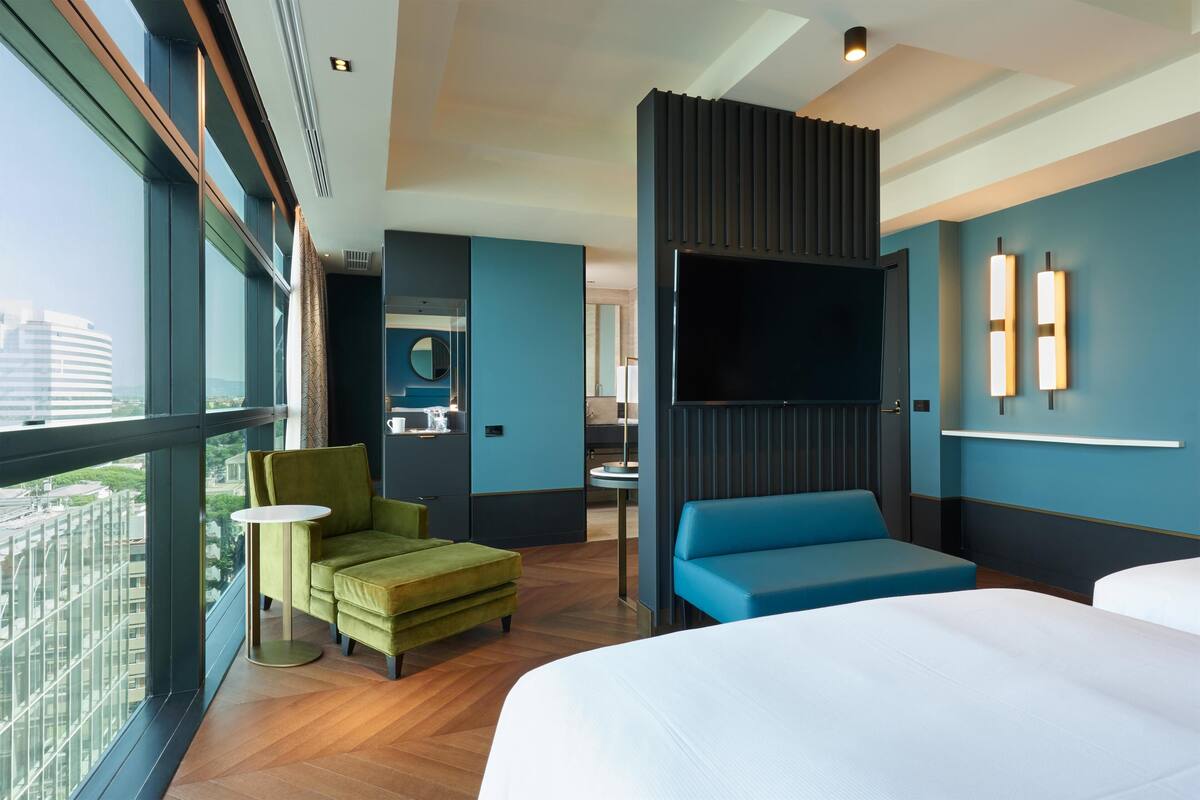 A modern hotel room with a large bed, a sofa, a TV, and a view of the city.