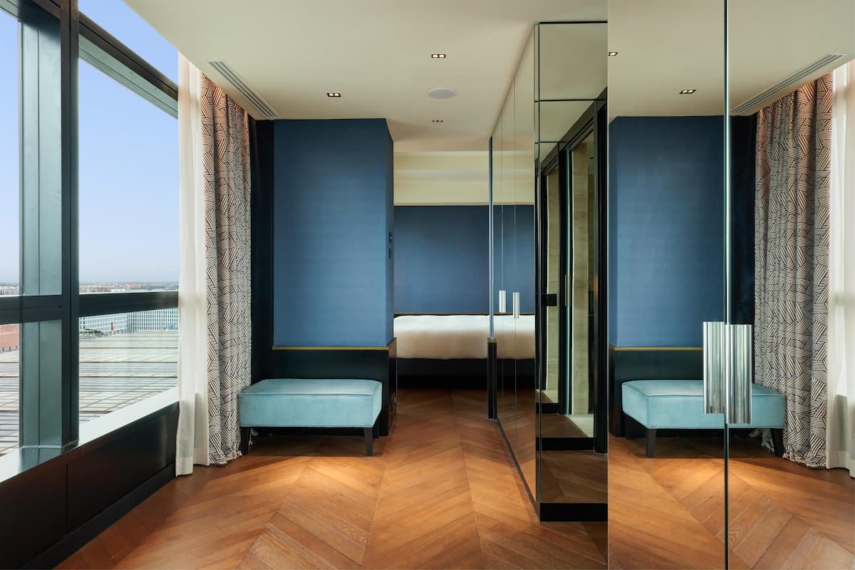 A modern hotel room with a large window, a bed, a bench, and a mirror.