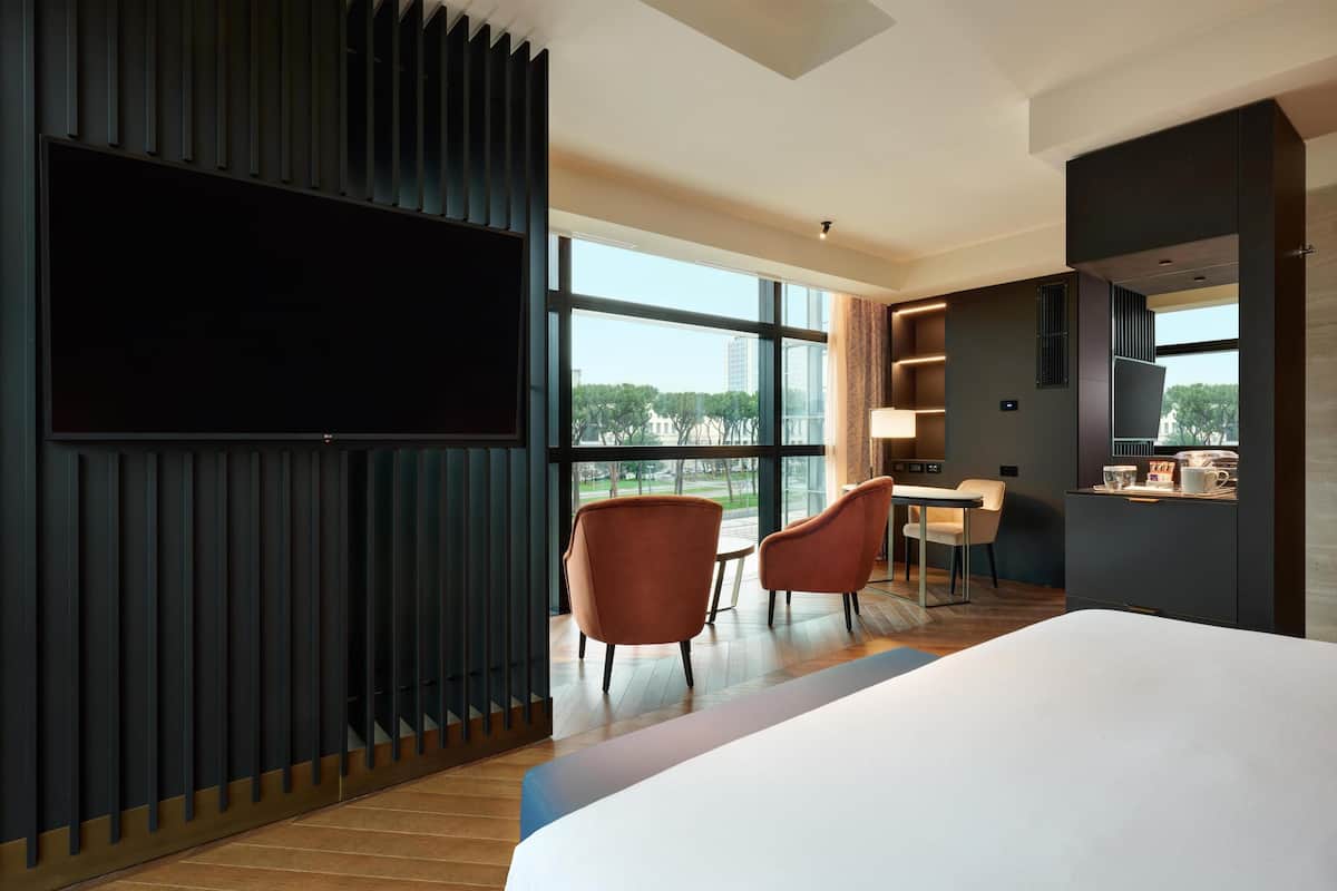 A modern hotel room with a large bed, a TV mounted on the wall, a desk with chairs, and a view of the city.