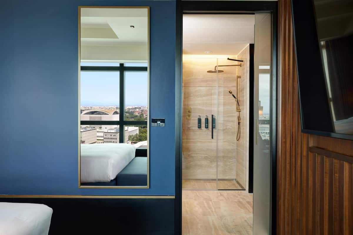 A modern hotel room with a large window, a bed, a shower area, and a television.