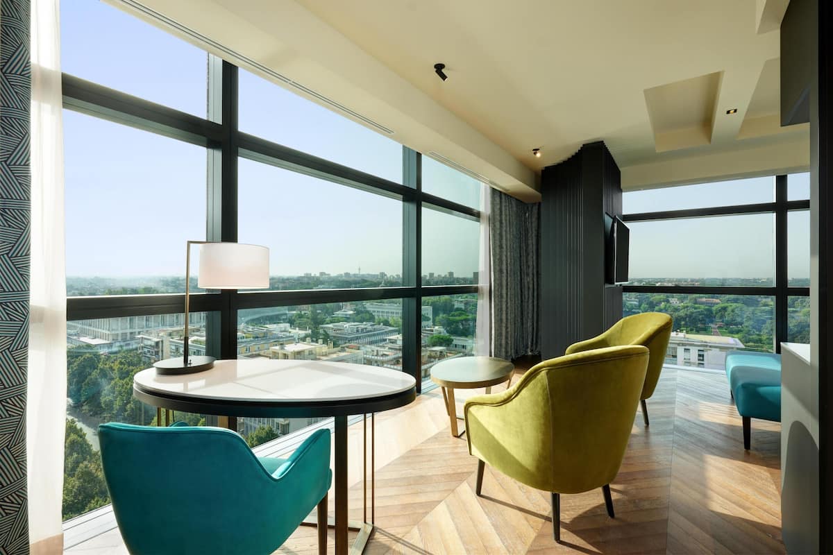 A modern hotel room with large windows, a round table, two chairs, a lamp, and a TV.