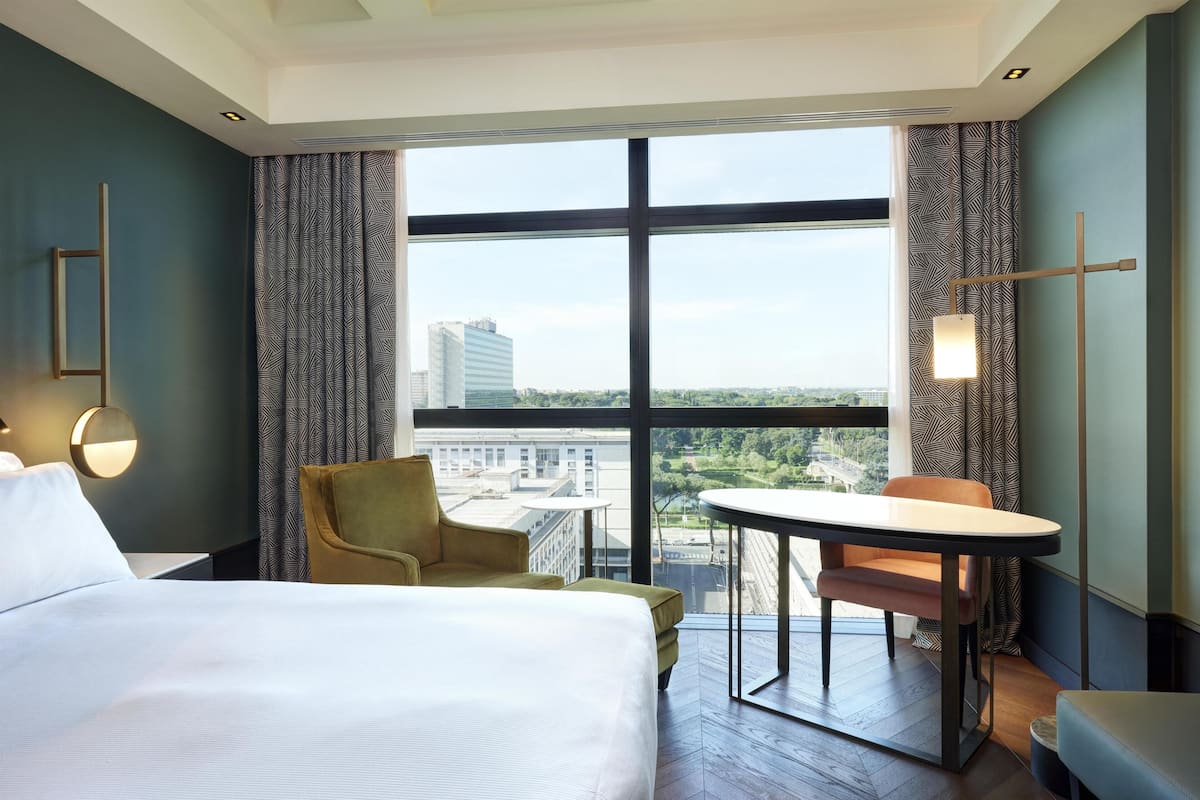 A hotel room with a large window, a bed, a chair, a table, and a lamp.