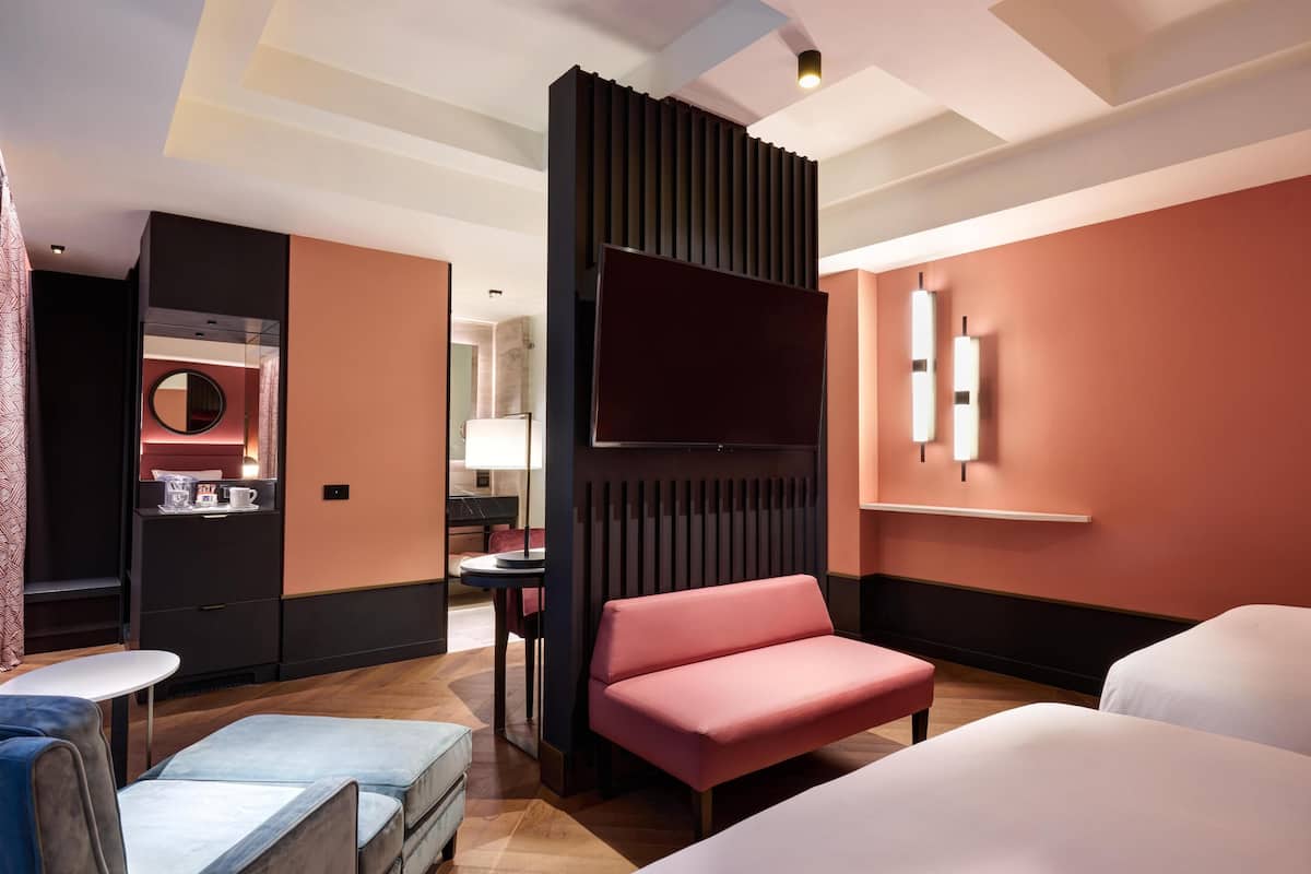 A hotel room with a bed, a sofa, a TV, a desk, and a bathroom visible in the background.