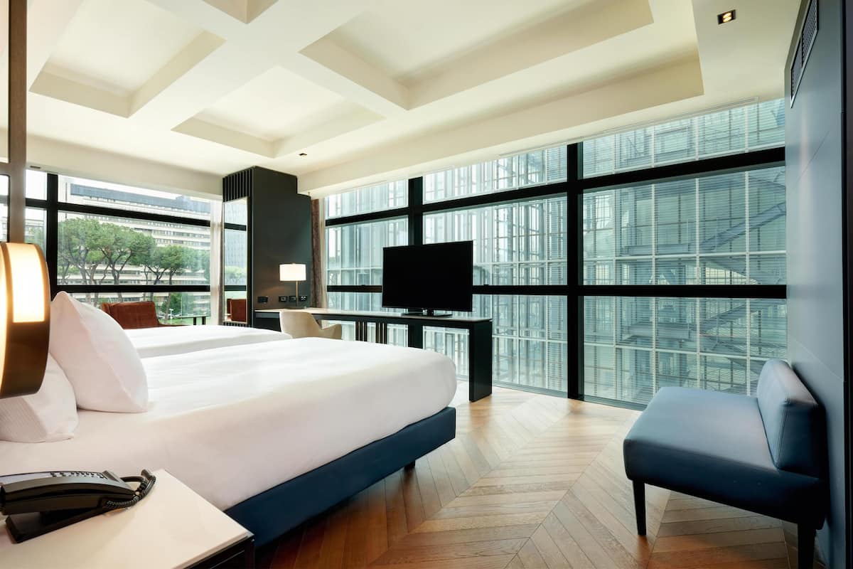 A modern hotel room with a large bed, a desk with a flat-screen TV, a chair, and large windows offering a city view.