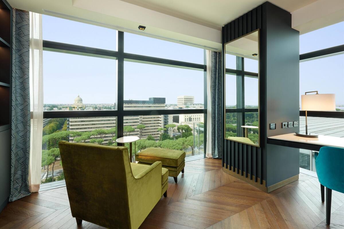 A modern hotel room with large windows, a sofa, a small table, and a desk with a lamp.