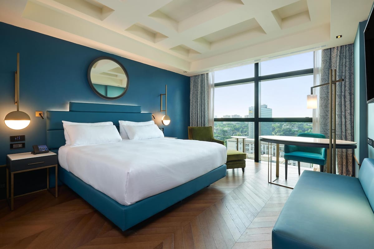A hotel room with a large bed, a round mirror, a desk, a chair, and a balcony with a table and chairs.