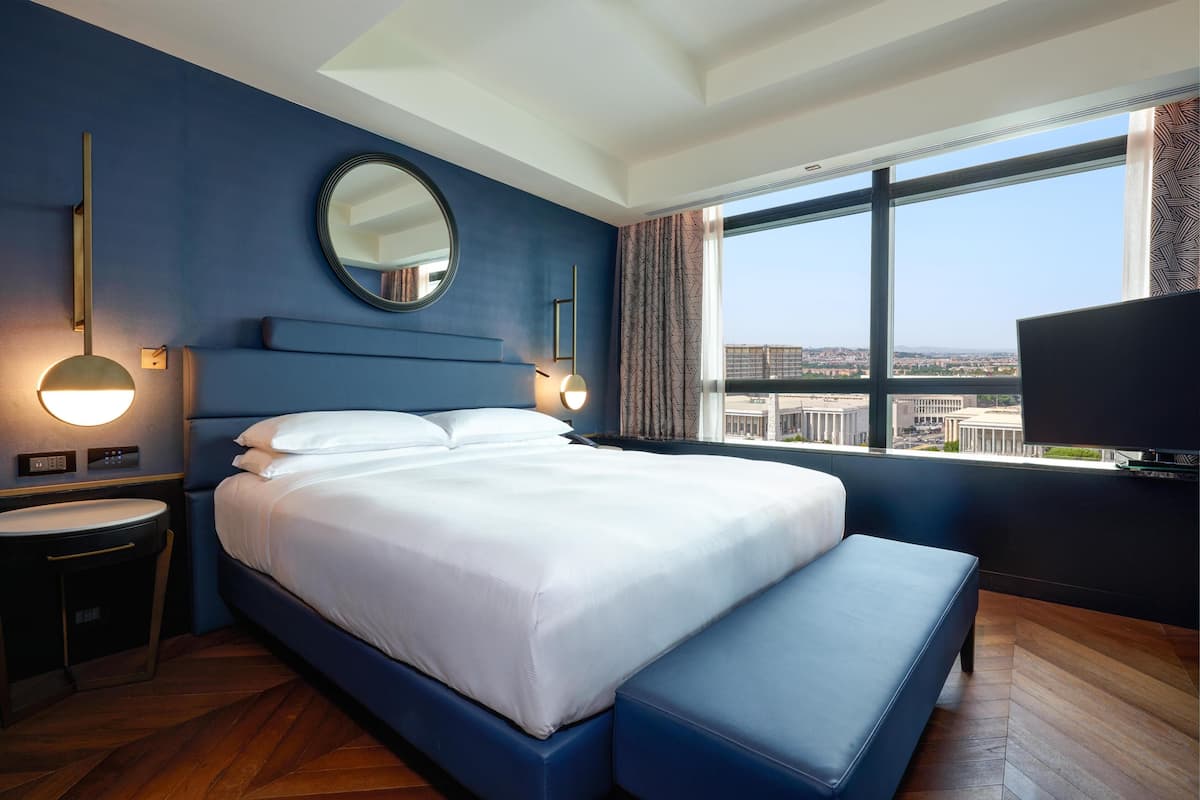 A hotel room with a large bed, a round mirror, a television, and a view of the city through a large window.