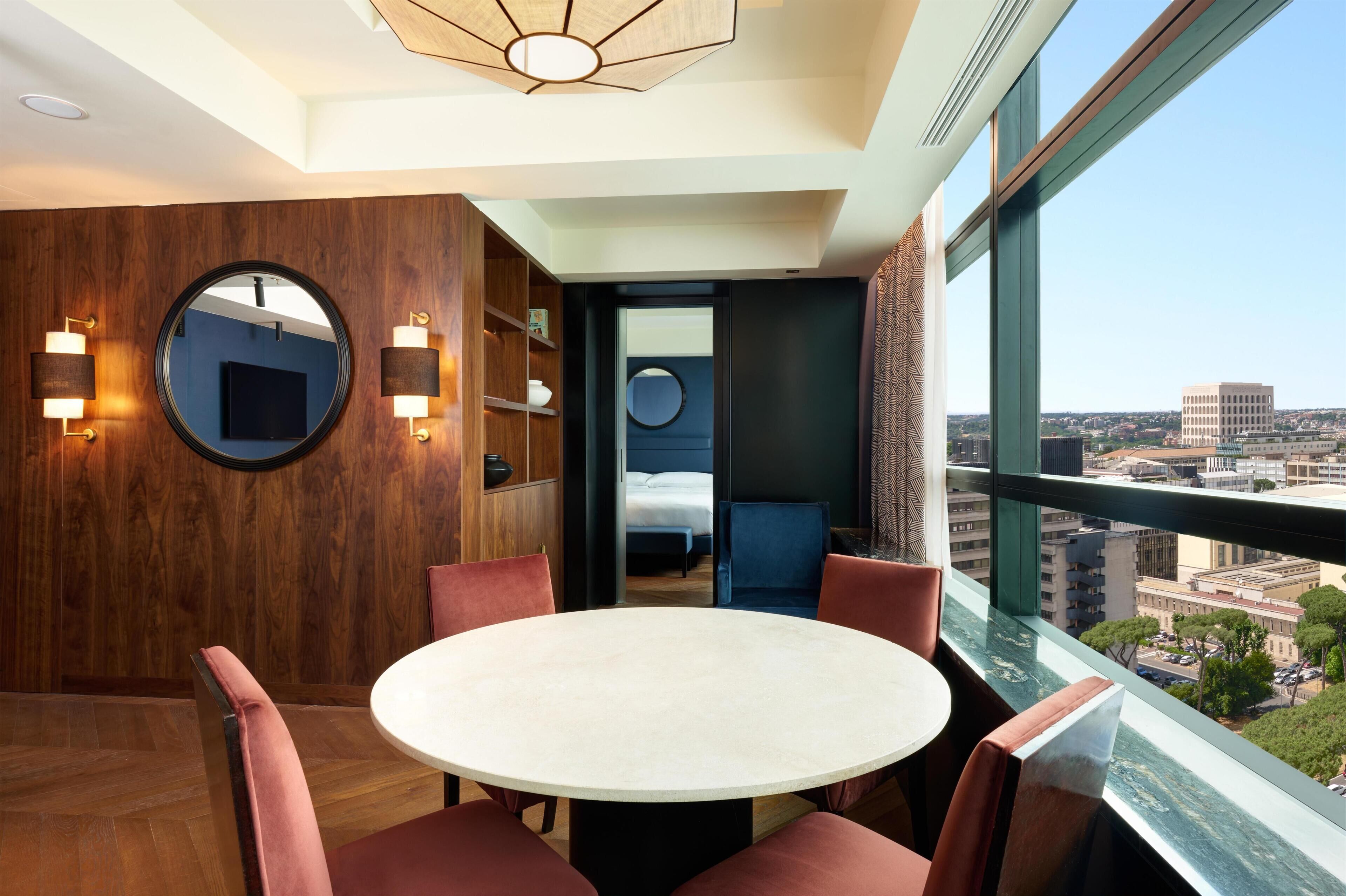 A modern hotel room with a round table, red chairs, a large window with a city view, and a wooden wall with a mirror and wall sconces.