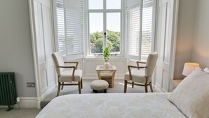 Ein Schlafzimmer mit einem großen Bett, zwei Stühlen, einem kleinen Tisch mit einer Vase und Blick durch ein Fenster mit Rollläden.