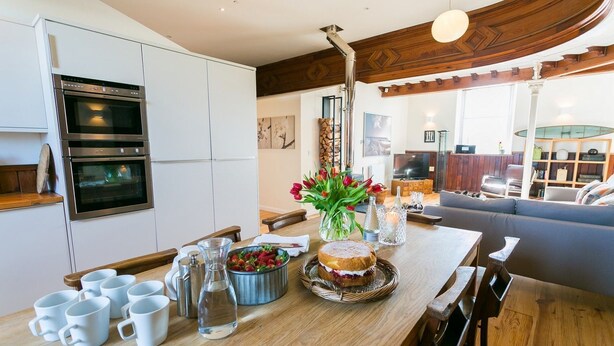 A modern kitchen with built-in ovens, a wooden dining table with a vase of flowers, and a cozy seating area with a sofa and a TV. - Cribinau (Ty Croes)
