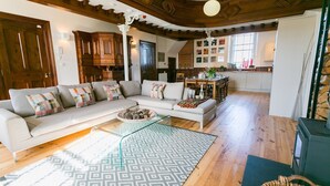 A spacious living room with a large sectional sofa, a glass coffee table, and a patterned rug. There is a kitchen area in the background with wooden cabinets and a dining table. - Cribinau (Ty Croes)