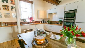 A modern kitchen with white cabinets, a wooden countertop, and a dining table set with a cake and tulips. - Cribinau (Ty Croes)