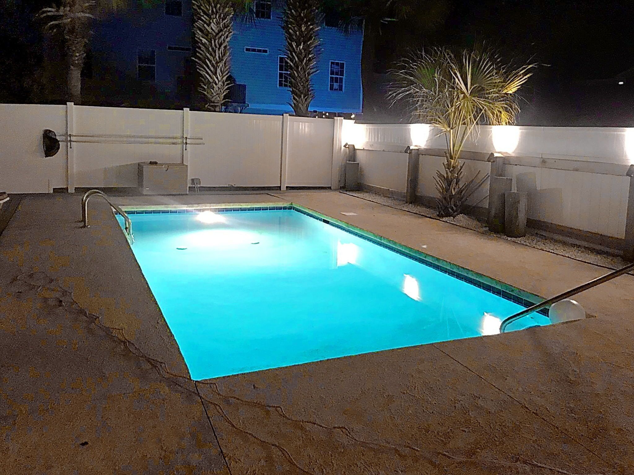A rectangular swimming pool with a clear blue water surface, surrounded by a white fence and palm trees.