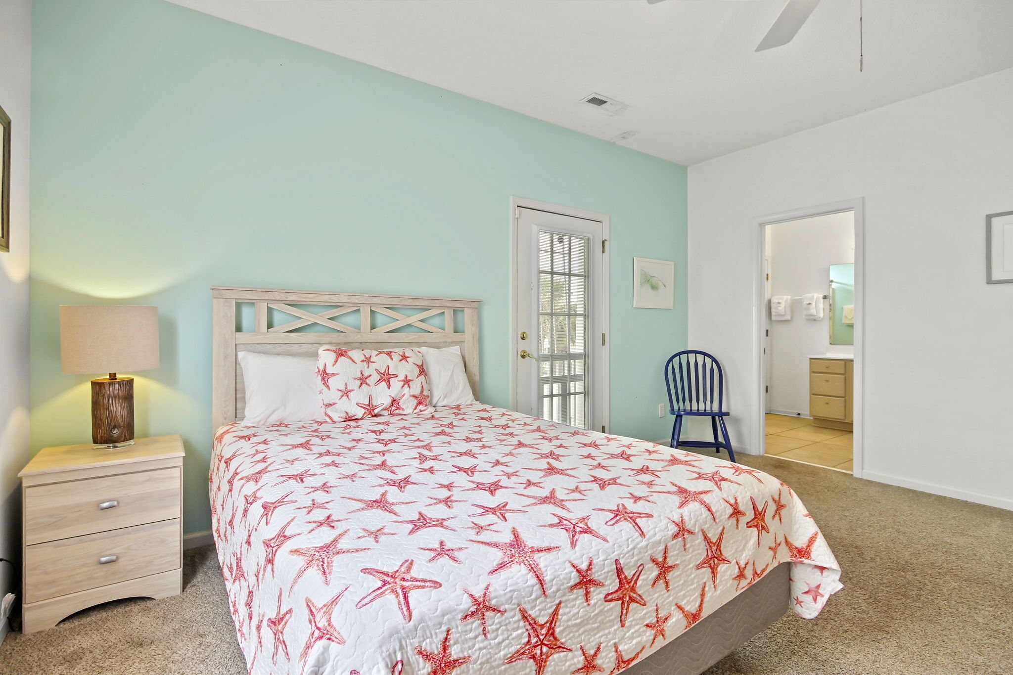 A bedroom with a bed, nightstand, lamp, chair, and a door leading to another room.