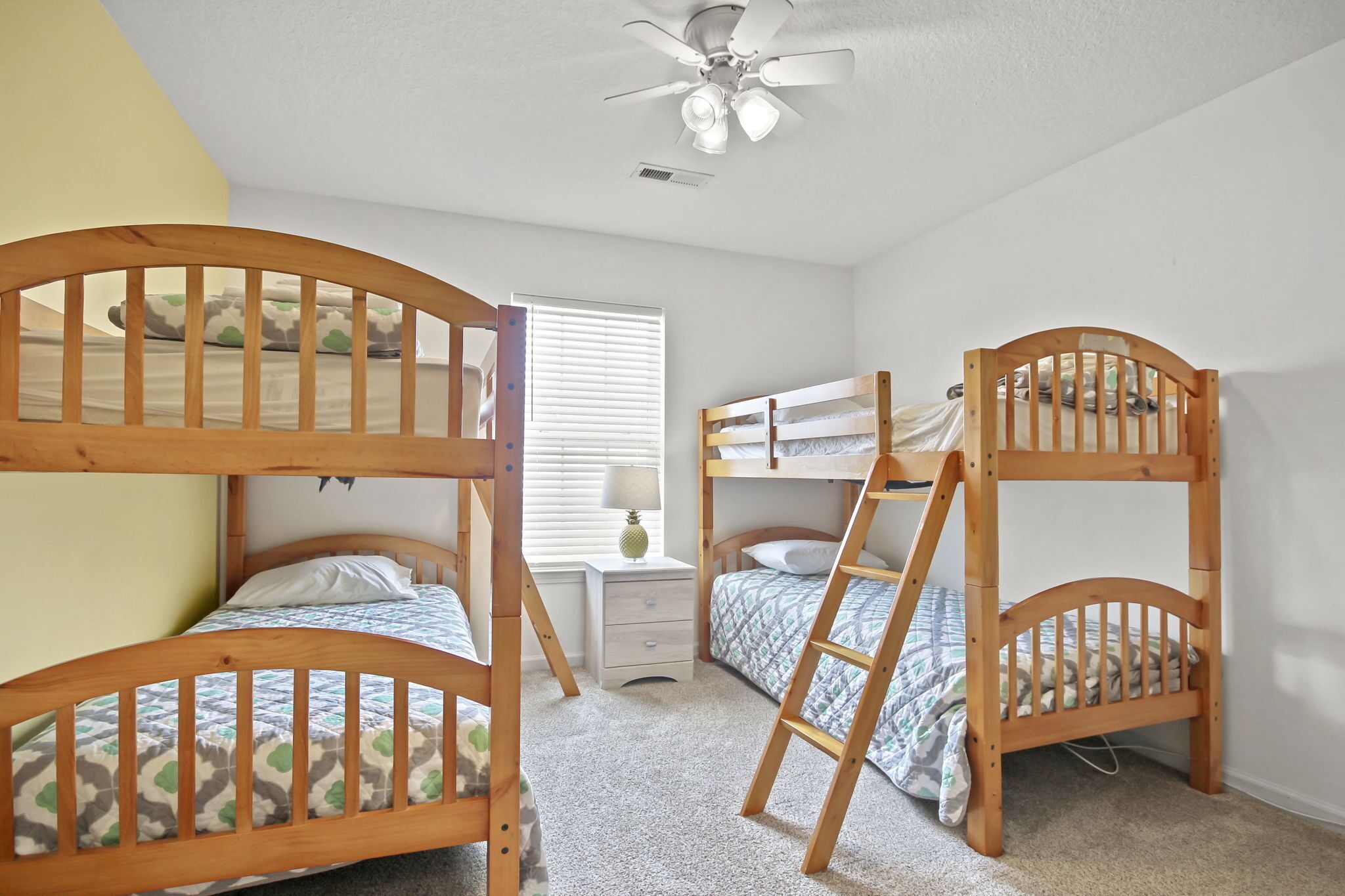 A bedroom with three bunk beds, a ceiling fan, a window with blinds, and a bedside table with a lamp.