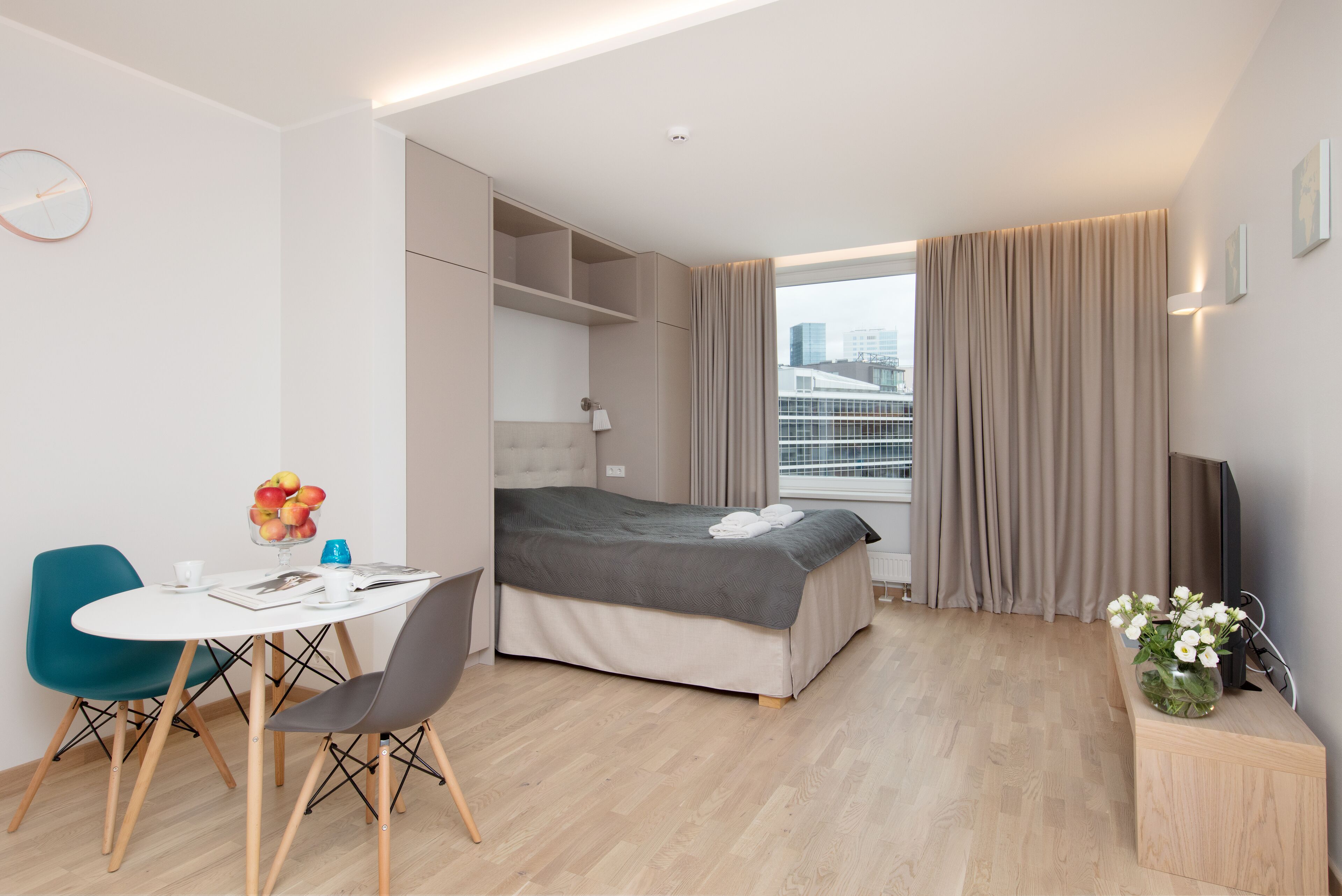Une chambre d’hôtel moderne comprenant un lit, un coin repas avec une table et des chaises, une petite fenêtre avec des rideaux et une vue sur la ville.