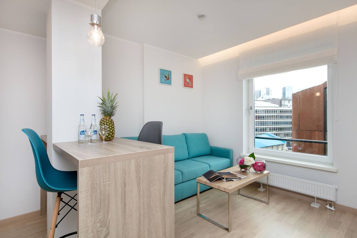 Ein modernes Wohnzimmer mit einem Holzesstisch, Türkis-Stühlen, einem Türkis-Sofa, einem Couchtisch mit Zeitschrift und Blumen und Blick auf die Stadt durch ein Fenster.