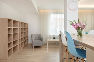 Een moderne kamer met een houten boekenkast, een grijze fauteuil, een eettafel met blauwe stoelen, en een bloevas.