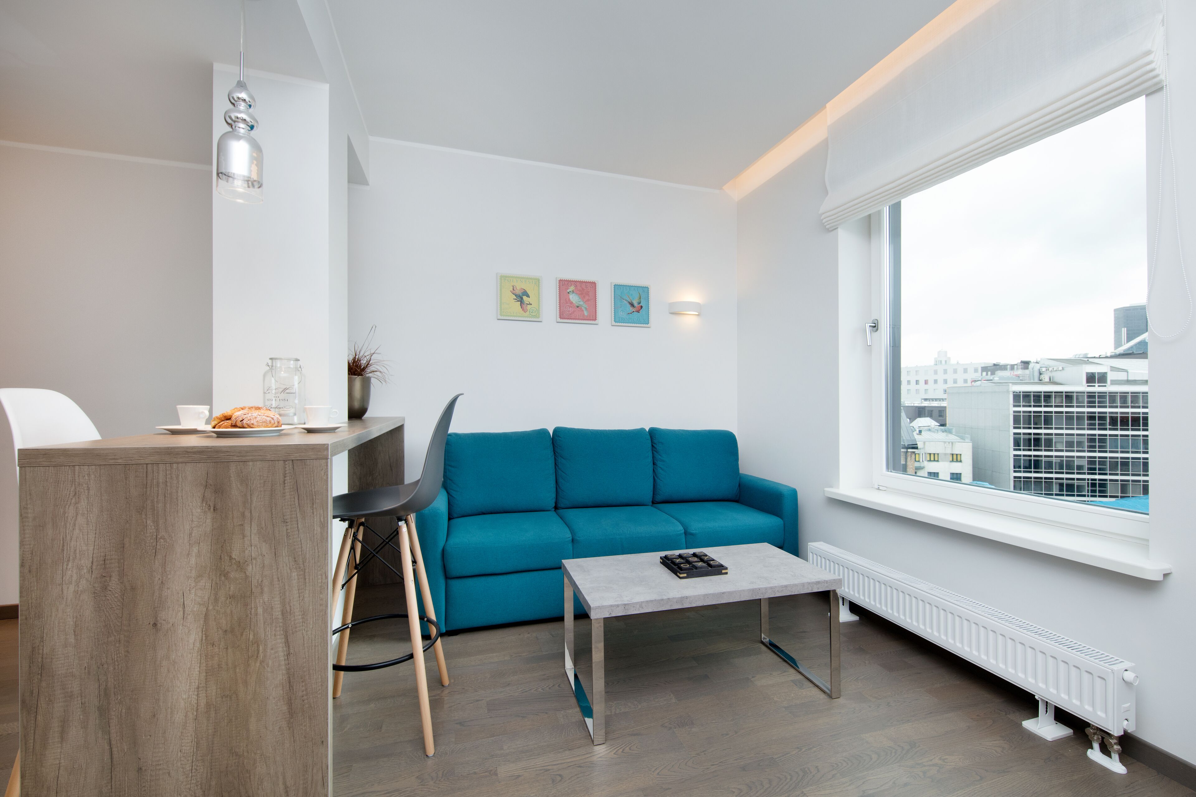 A modern living room with a teal sofa, a wooden coffee table, a white radiator, and a city view through a large window.