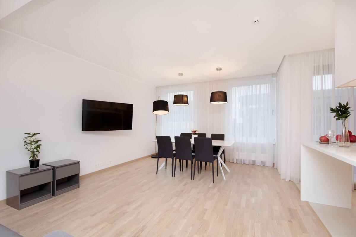 Ein modernes Wohnzimmer mit Flachbildfernseher, einem Essbereich mit Stühlen, einem kleinen Tisch mit einer Vase und einer Pflanze.