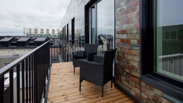 Un balcon doté de meubles en osier, d’un plancher en bois et d’un mur en briques agrémenté de grandes fenêtres.