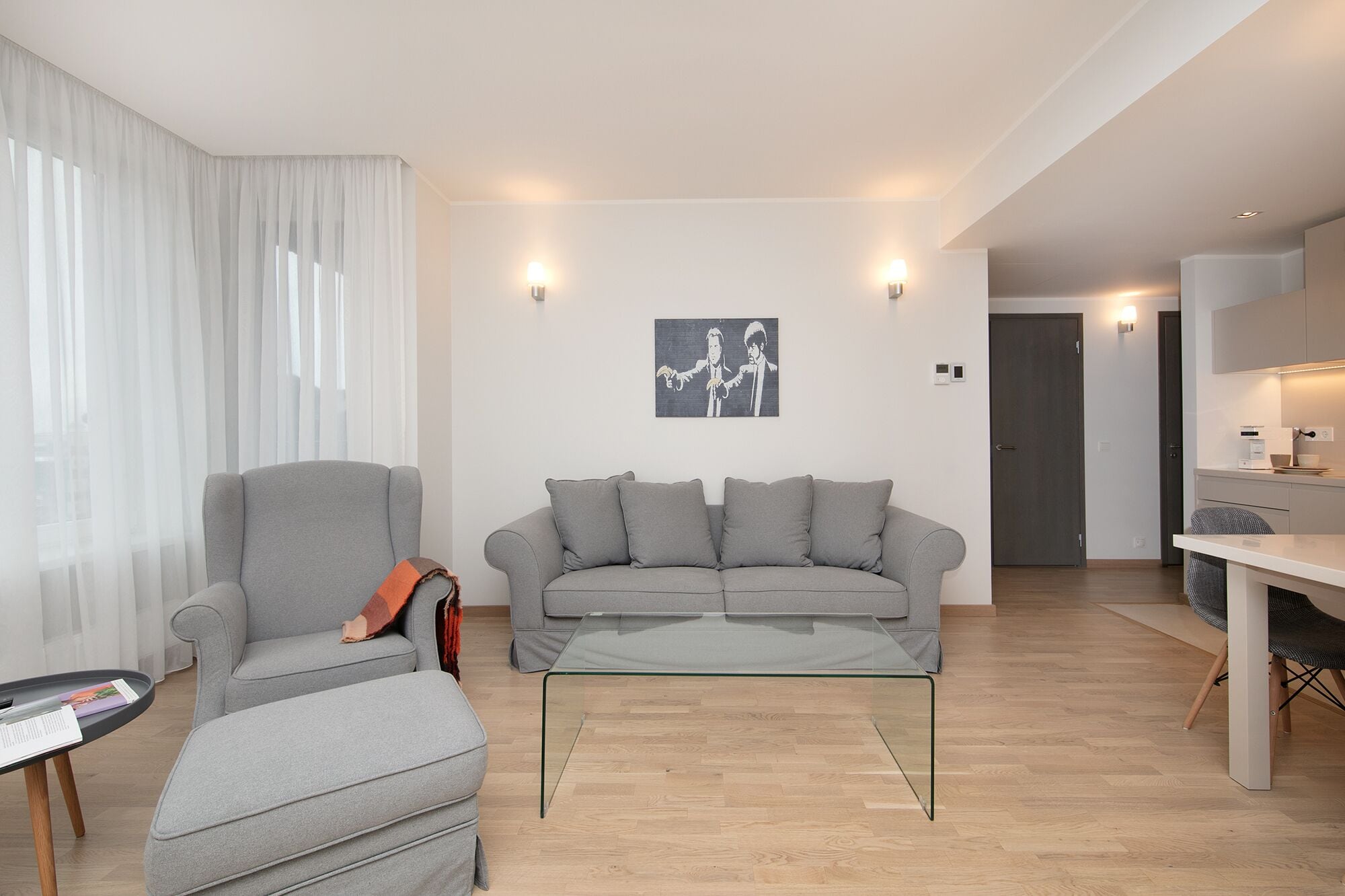 A modern living room with a grey sofa, a grey armchair, and a glass coffee table. There is a painting on the wall and a door leading to another room.
