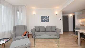 A modern living room with a grey sofa, a grey armchair, and a glass coffee table. There is a painting on the wall and a door leading to another room.