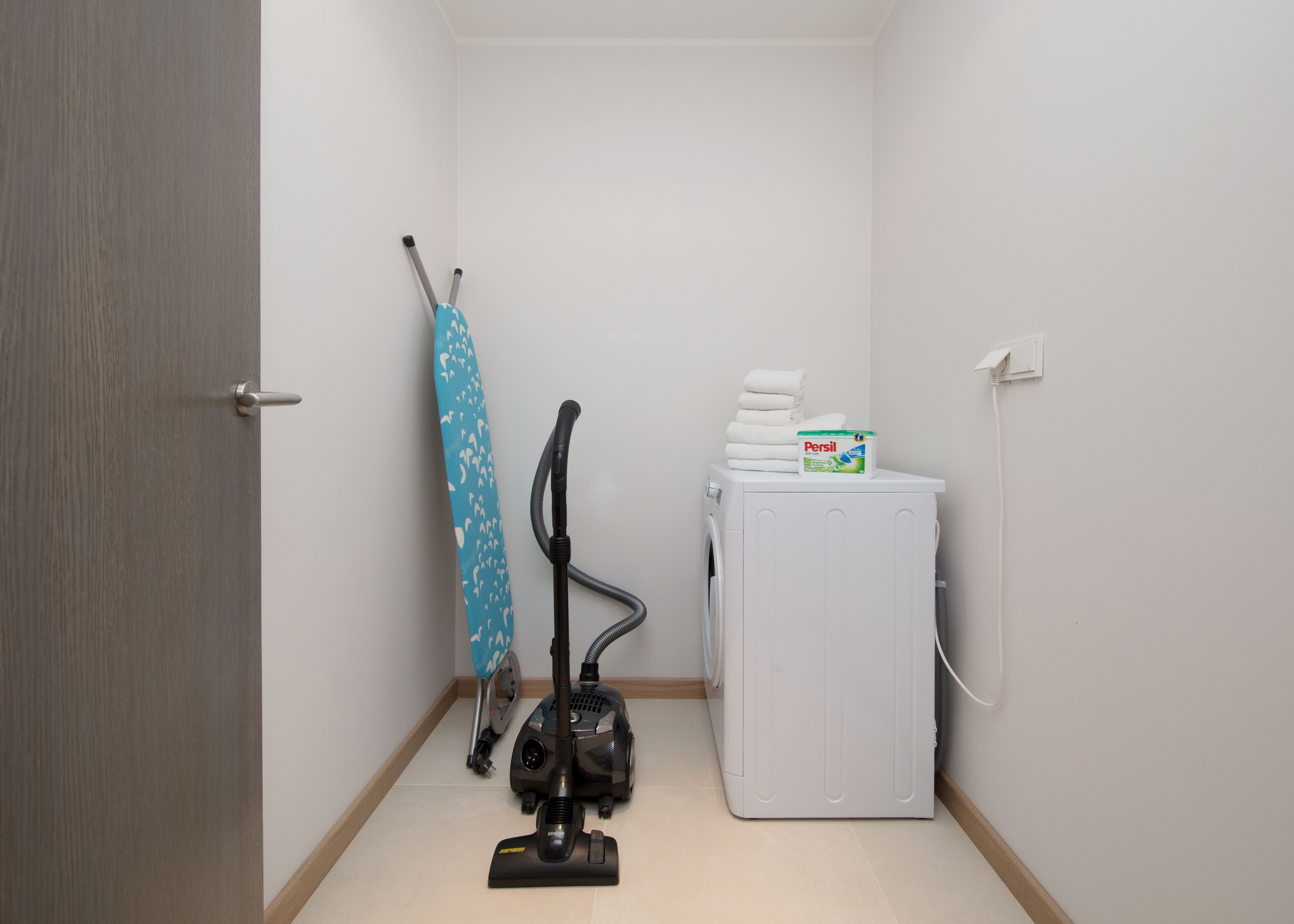 A laundry room with a washing machine, a vacuum cleaner, and a stack of white towels.