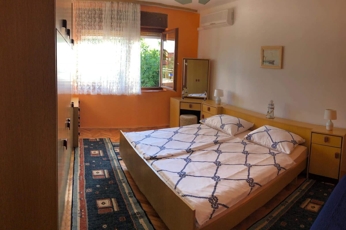 A bedroom with a bed, a nightstand, a mirror, and a window with curtains.
