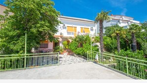 A building with a white facade, yellow shutters, and a balcony. There are trees and a palm tree in the foreground. - Apartments Andrea (Crikvenica)