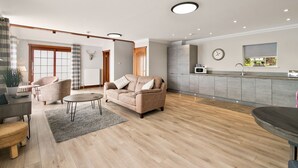 A spacious living room with a beige sofa, a coffee table, and a dining area with a table and chairs. There is a kitchen area with a microwave and a window. - Creinch Lodge-Sleeps 2 - sleeps 2 guests  in 1 bedroom (Alexandria)