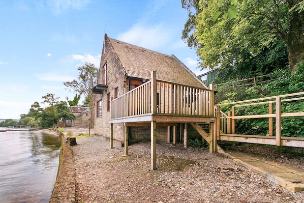 Une construction rustique en bois, dotée d’un toit en pente, d’une terrasse en bois et de fondations en pierre, située près d’une étendue d’eau.