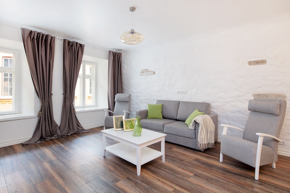 Ein modernes Wohnzimmer mit grauem Sofa, weißem Couchtisch und grauem Sessel. Ein Fenster mit Vorhängen, eine Pendelleuchte und eine weiße Wand mit einer kleinen dekorativen Platte.