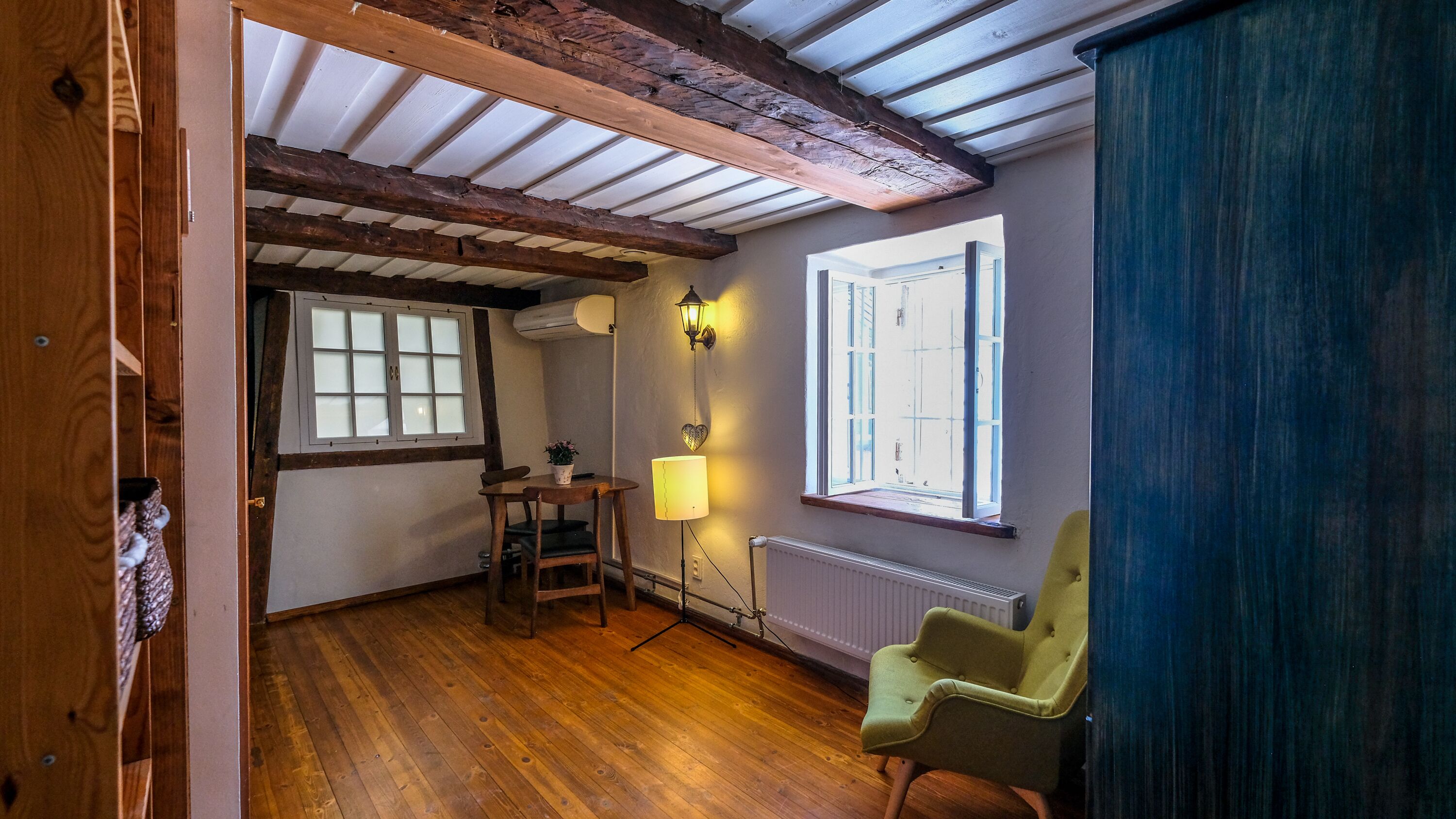 Een kamer met houten balken, een raam met zonwering, een tafel met een lamp en een stoel.