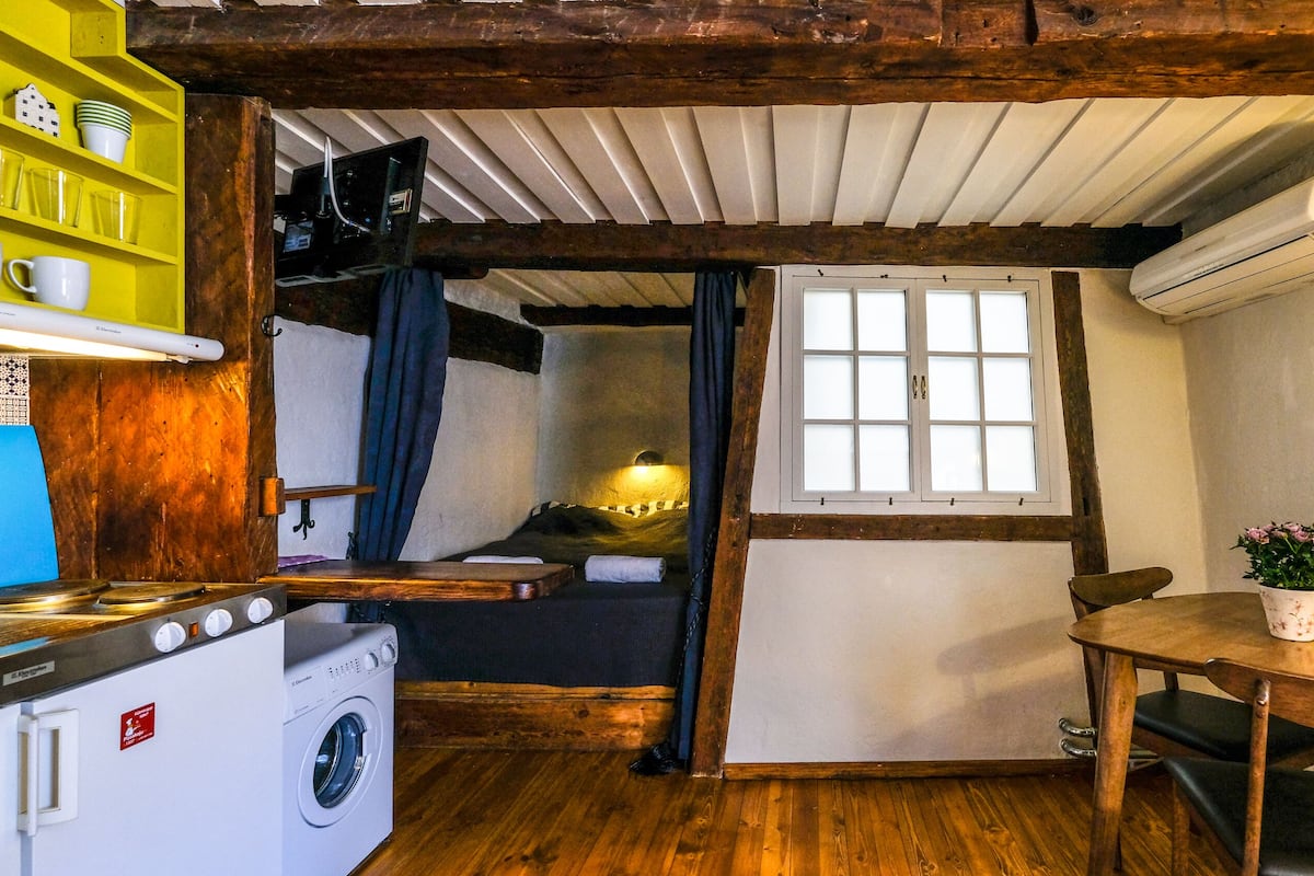 Eine kleine Küche mit Holzbalken, einer Waschmaschine, einem Esstisch und einem fenster mit Vorhängen.